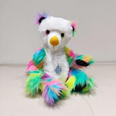A handmade bear by Brierley Bears. White body and head with bright multicoloured lims and ears and black paws. Seated, looking forward.